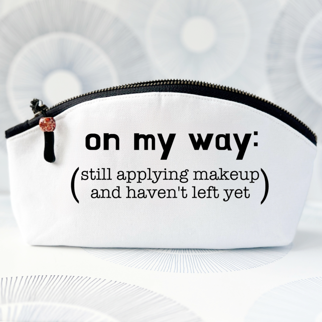 white canvas zip bag with a curvy top, a black zipper and the words, on my way: still applying makeup and haven't left yet.
