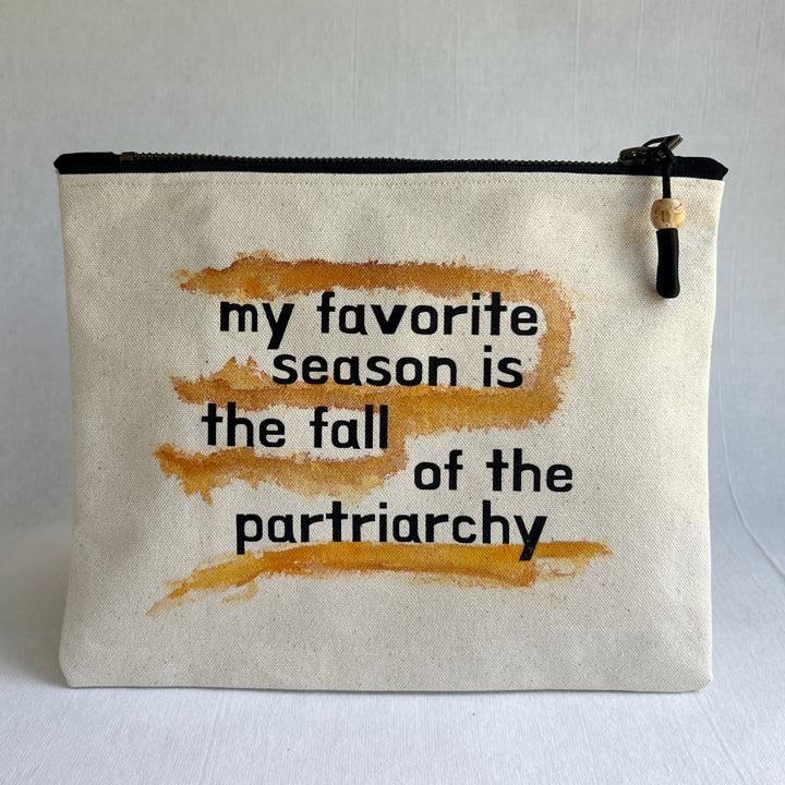 An off-white canvas zipper pouch with a black zipper and a wooden tassel. The front features a black printed text reading 'my favorite season is the fall of the partriarchy' overlaid on top of three horizontal orange-brown watercolor brushstrokes.