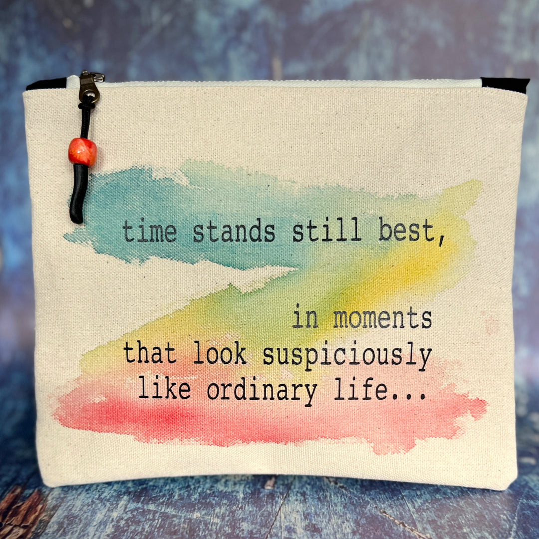 rectangular canvas bag with a zipper pull, in linen-colored duck cloth canvas. Watercolor paint in blues, yellows and reds is the background for the words, time stands still best, in moments that look suspiciously like ordinary life...