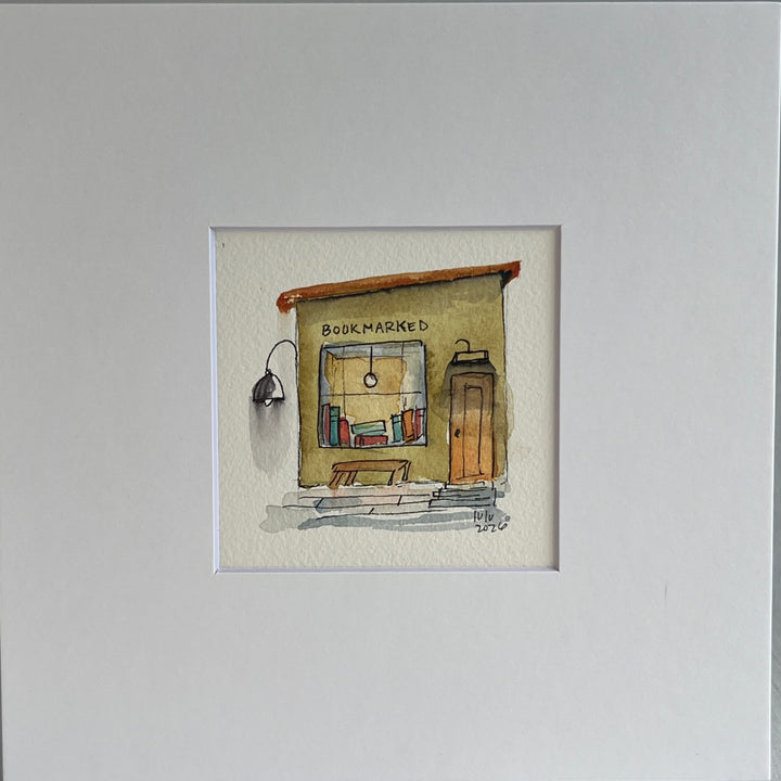A matted watercolor painting of a cozy bookshop storefront with the word 'BOOKMARKED' above a window filled with books, a bench, a door, and a hanging lamp, shown on a white mat and white frame.