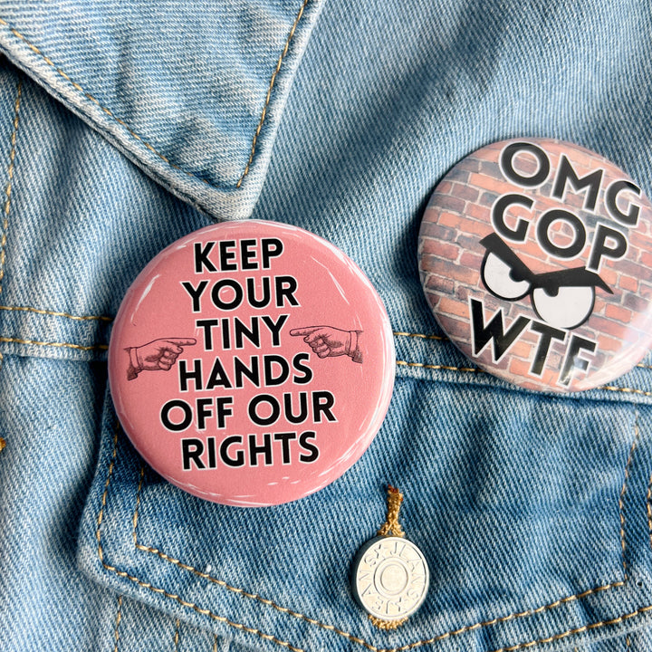 round pinback button with a pink background and the words, "Keep your tiny hands off our rights." A pair of hands in black point at the words. Pinned on a jean jacket for display. A second button has the acronyms OGM GOP WTF with frowning eyes, on a red brick background.
