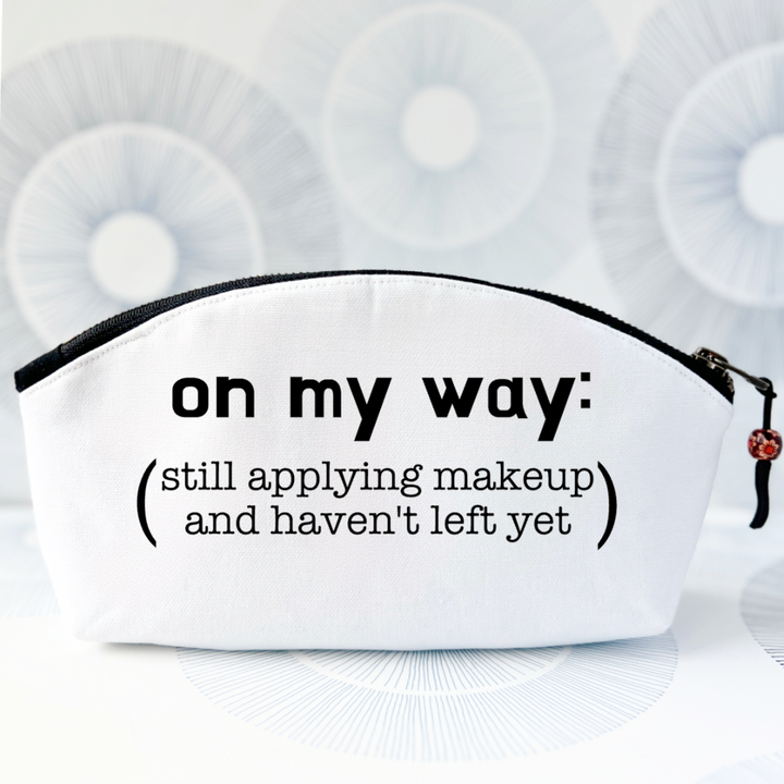 white canvas zip bag with a curvy top, a black zipper and the words, on my way: still applying makeup and haven't left yet.