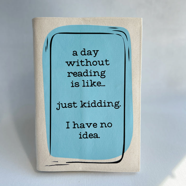 A canvas, reusable book cover in linen, with the words, 'a day without reading is like.. just kidding. I have no idea. ' in black lettering, placed on a rectangular light blue box design.
