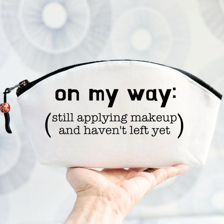 white canvas zip bag with a curvy top, a black zipper and the words, on my way: still applying makeup and haven't left yet.