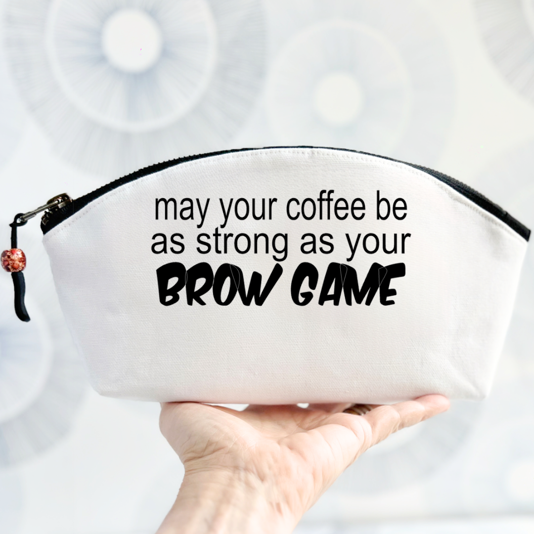 white canvas bag with a curvy top, a black zipper and the words, may your coffee be as strong as your brow game.