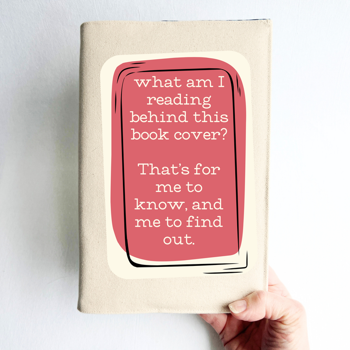 A hand holding a book covered with a reusable canvas book cover in linen, with the words, 'what am I reading behind this book cover?  That's for me to know, and me to find out' in cream lettering, placed on a rectangular coral box design.