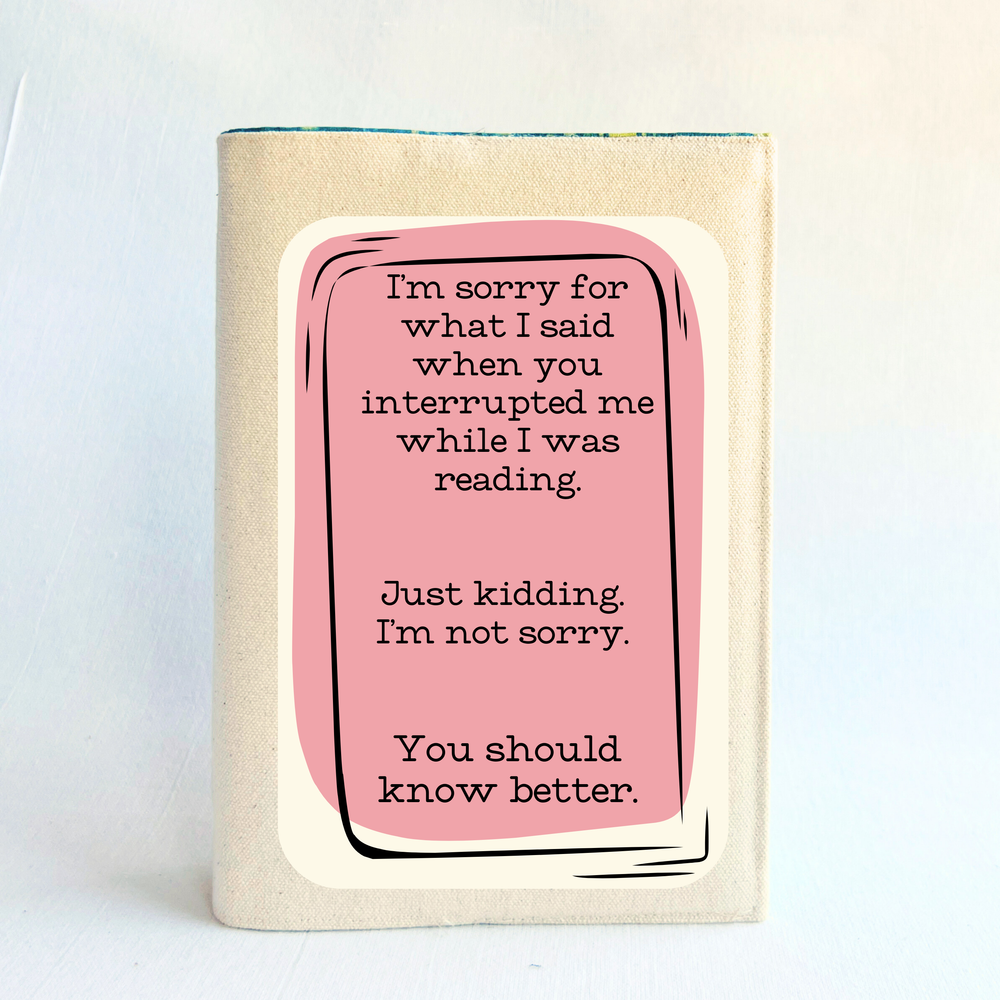 a book covered with a linen-color reusable, readjustable book cover.  A pink square background has the words, 'I'm sorry for what I said when you interrupted me while I was reading.  Just kidding. I'm not sorry. You should know better.' in black lettering.