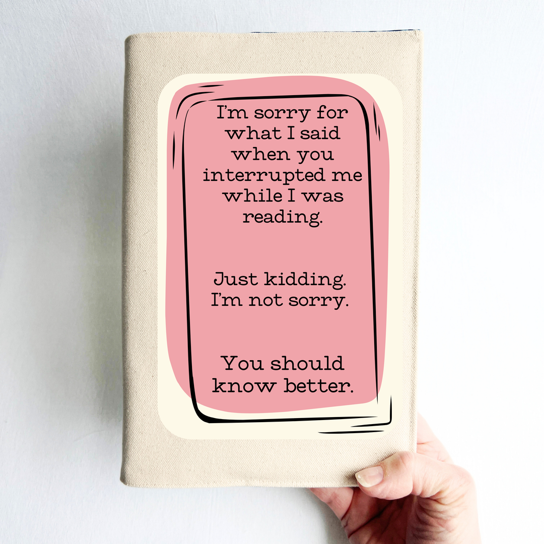 a hand holding a book covered with a linen-color reusable, readjustable book cover.  A pink square background has the words, 'I'm sorry for what I said when you interrupted me while I was reading.  Just kidding. I'm not sorry. You should know better.' in black lettering.