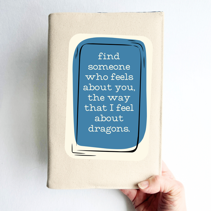 a hand holding a book covered in linen-color reusable canvas adjustable book cover. A blue square is the background for the words, 'find someone who feels about you, the way that I feel about dragons.' in white lettering.