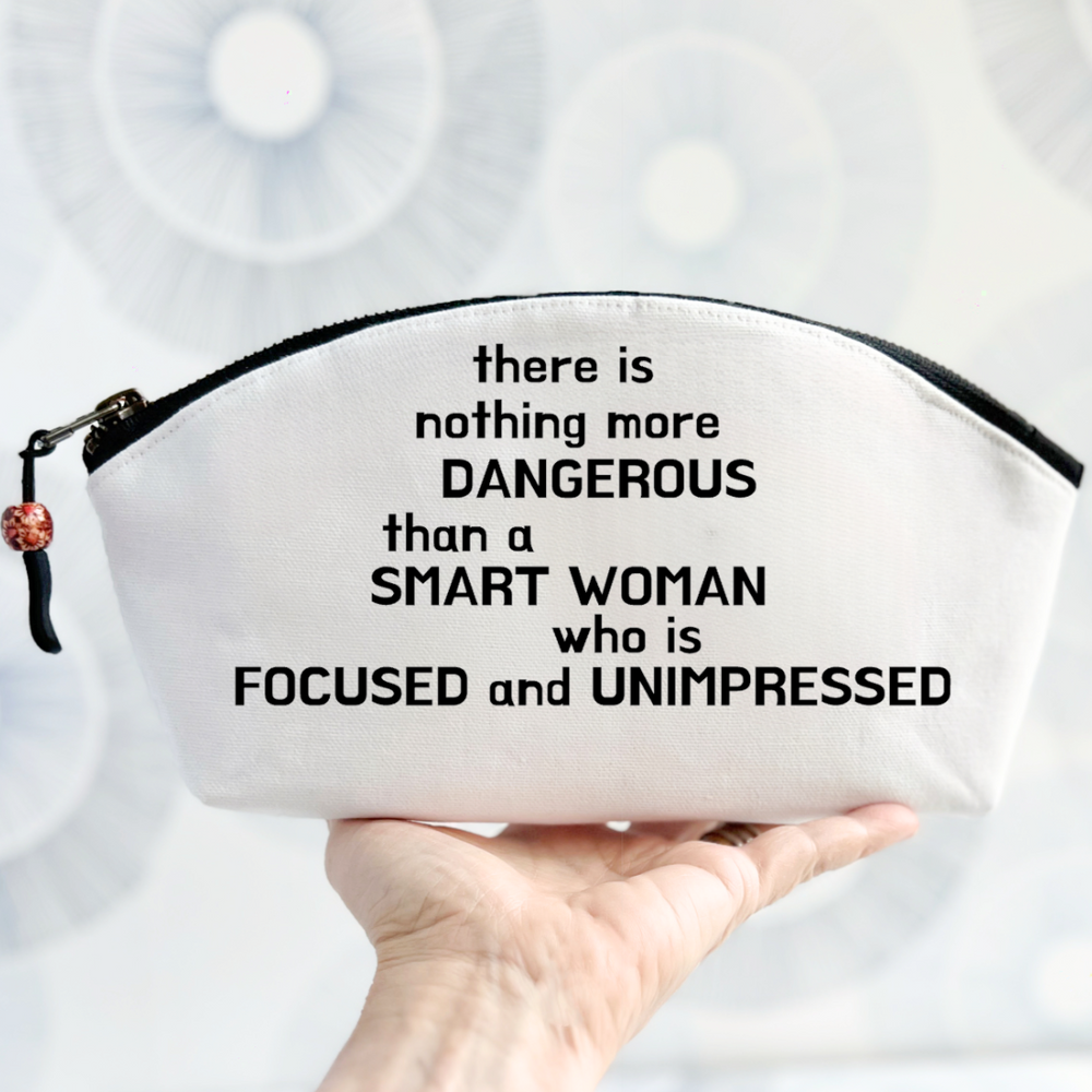 white canvas bag with curvy top, black zipper and the words, there is nothing more dangerous than a smart woman who is focused and unimpressed