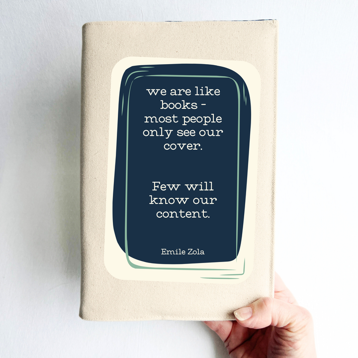 a hand holding a book covered with a linen-colored canvas reusable book cover.  A rectangular design in navy tops the cover, with the words, 'we are like books - most people only see our cover.  Few will know our content.' a quote by Emile Zola, done in white lettering.