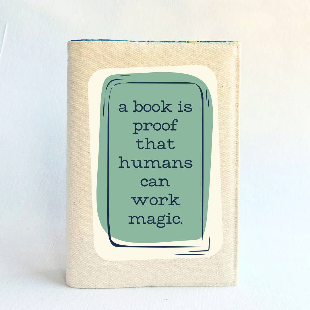 a book covered with a linen-colored canvas reusable book cover. A rectangular design in sage green is featured in the center of the cover, with the words, 'a book is proof that humans can work magic.'