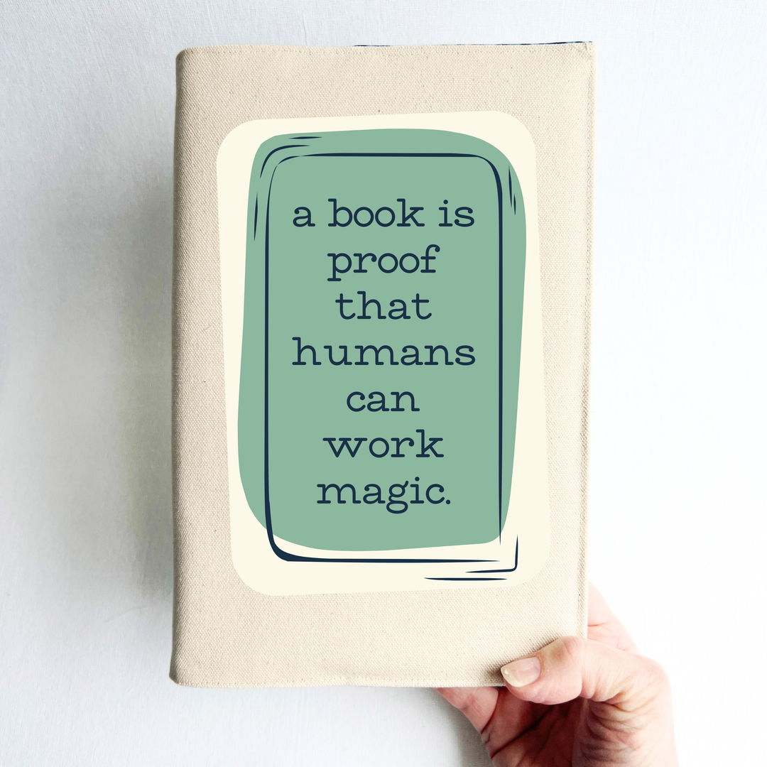 a hand holding a book covered with a linen-colored canvas reusable book cover. A rectangular design in sage green is featured in the center of the cover, with the words, 'a book is proof that humans can work magic.'