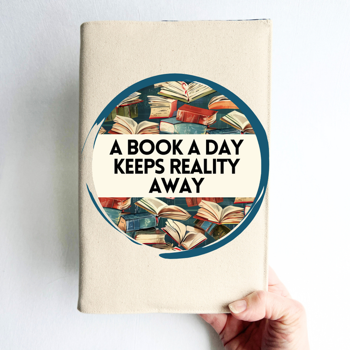 a hand holding a book covered with a linen-colored canvas reusable book cover. A circular design with painted books is featured in the center of the cover, with the words, 'a book a day keeps reality away.'