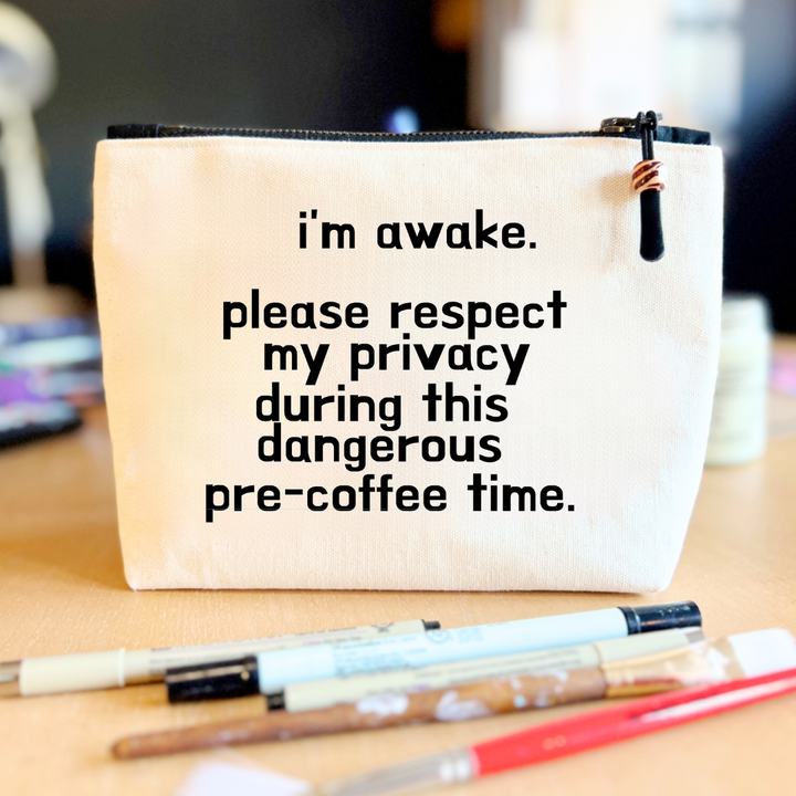 square linen-color canvas zip bag with a black zipper and the words, I'm awake. please respect my privacy during this dangerous pre-coffee time.