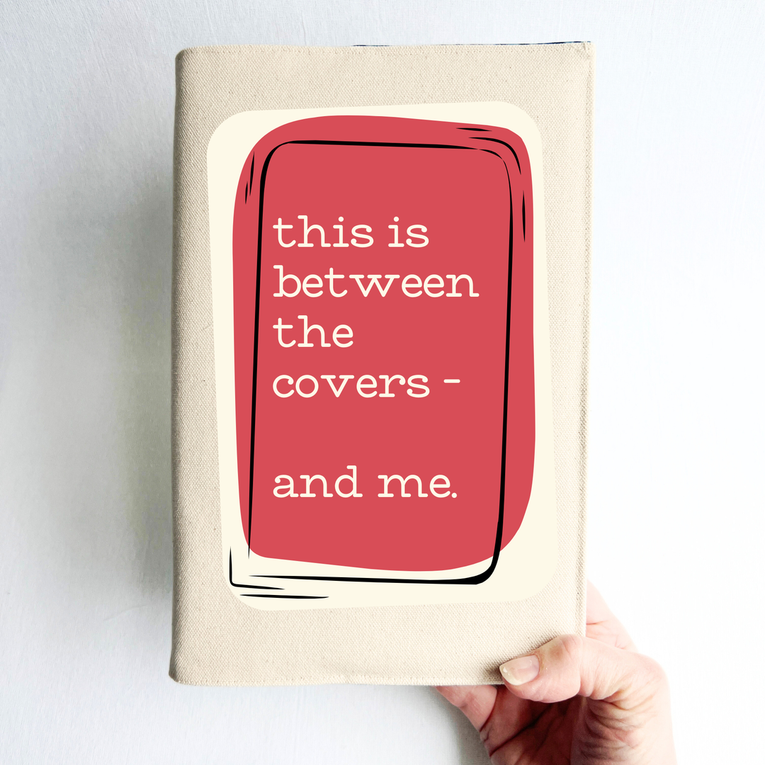 A hand holding a linen-colored canvas book cover with a rose and cream graphic cover that reads 'this is between the covers - and me.' against a plain light background.