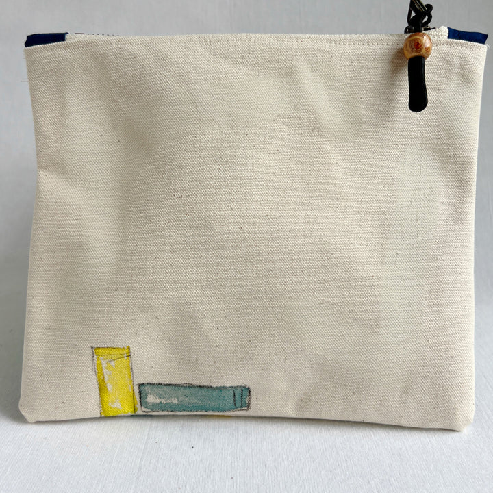The back side of a square canvas pouch in natural cream canvas with a watercolor-style illustration of colorful book spines along the bottom. The pouch has a top zipper with a black cord pull and a wooden bead accent.