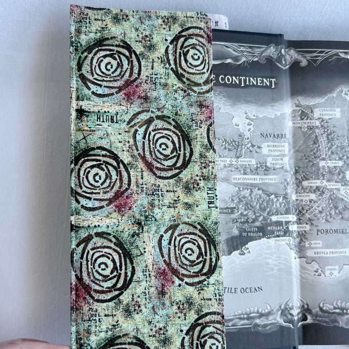 a book covered in a vibrant blue green fabric reusable  adjustable book cover.  Fabric has impressionist rose / concentric deep brown circles and the word ' TRUTH scattered on the pattern.