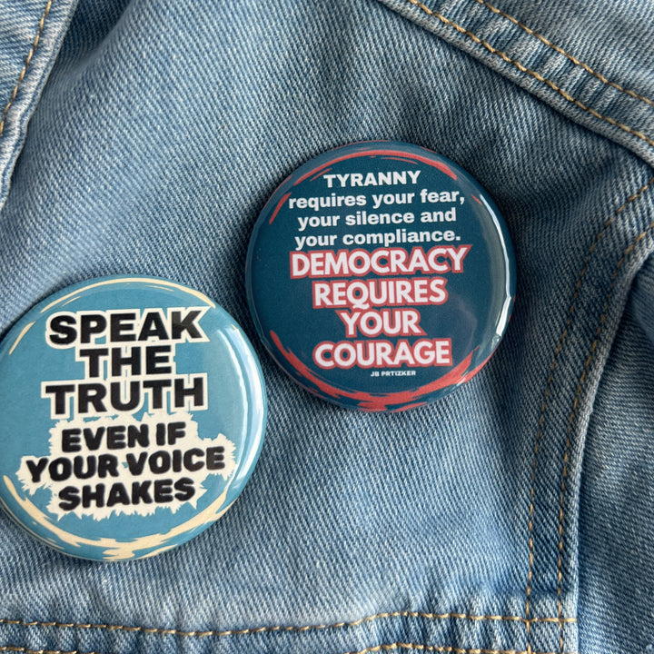 round pinback button with a blue background and the words, 'speak the truth even if your voice shakes' in black lettering bordered in white. Another pinback button in deep blue with the words, 'tyranny requires your fear, your silence and your compliance. Democracy requires your courage' in white lettering. both buttons are pinned to a jean jacket for display