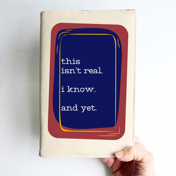 A person's hand holding a linen-colored canvas book cover with a graphic design on the cover. The design features a dark blue central rectangle with white text reading 'this isn't real. I know. And yet.'