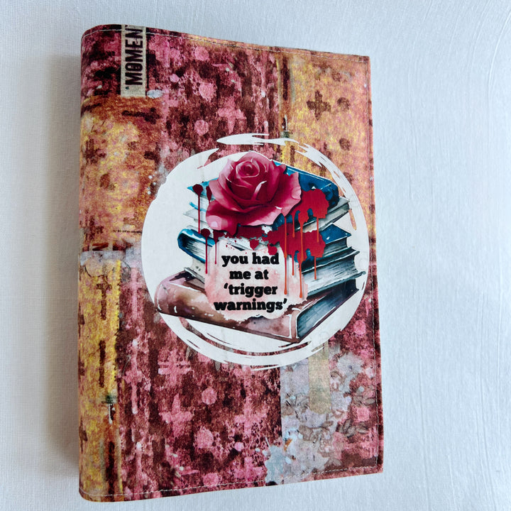 a book covered in a colorful fabric reusable book cover in colors of pinks and orange. A white circle on the front contains an original design with a stack of books, a rose, blood drips and the words, 'you had me at trigger warnings' finishes the look.  Meant for readers of dark romance.