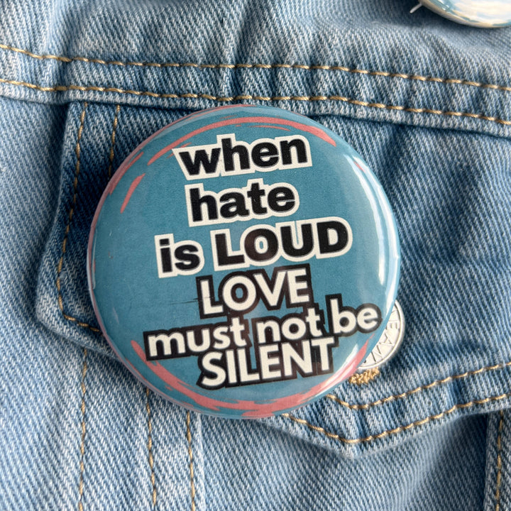 round pinback button on a blue background with the words, 'when hate is loud, love must not be silent' in black and white lettering and pinned to a jean jacket for display.