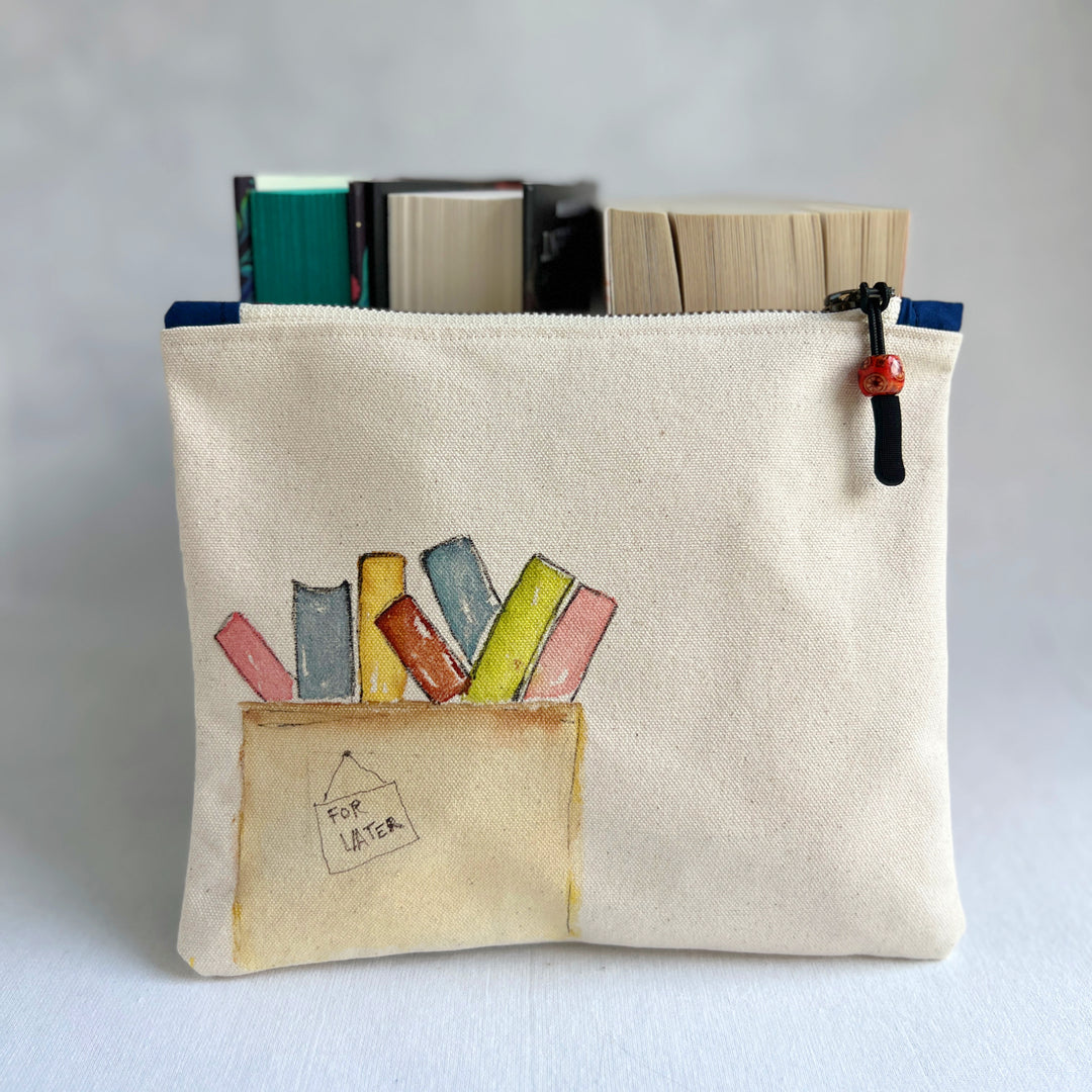 A canvas zippered pouch with a watercolor illustration of colorful books in a box labeled 'FOR LATER'. Several books are tucked inside the pouch, which is photographed against a light gray background.