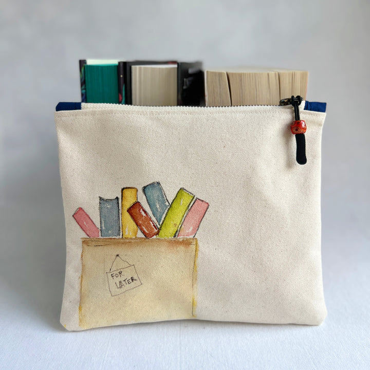 A canvas zippered pouch with a watercolor illustration of colorful books in a box labeled 'FOR LATER'. Several books are tucked inside the pouch, which is photographed against a light gray background.