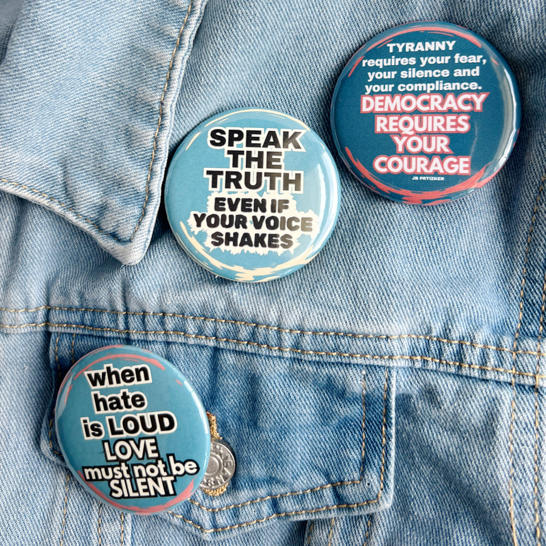 round pinback button with a blue background and the words, 'speak the truth even if your voice shakes' in black lettering bordered in white. Another pinback button in deep blue with the words, 'tyranny requires your fear, your silence and your compliance. Democracy requires your courage' in white lettering. A third button says, 'when hate is loud, love must not be silent' with a blue background and white lettering. All buttons are pinned to a jean jacket for displa