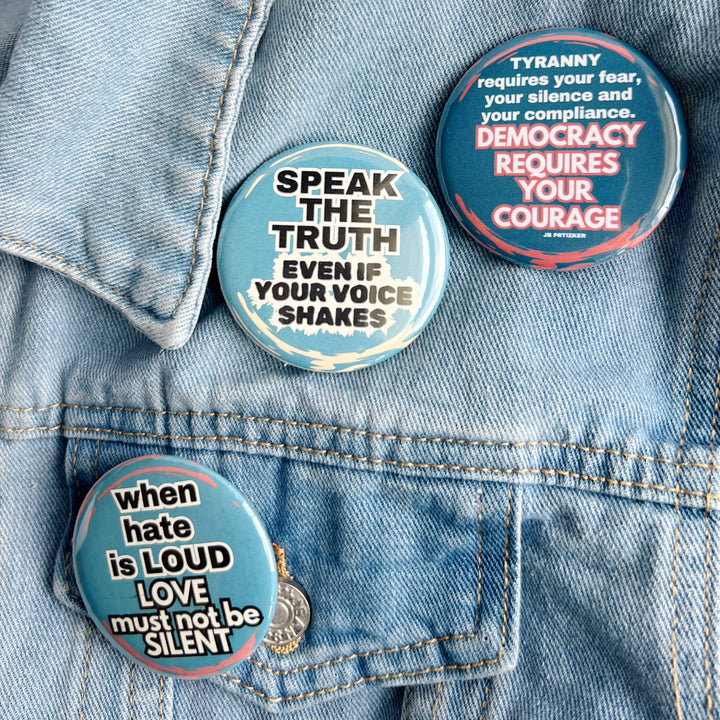 round pinback button with a blue background and the words, 'speak the truth even if your voice shakes' in black lettering bordered in white. Another pinback button in deep blue with the words, 'tyranny requires your fear, your silence and your compliance. Democracy requires your courage' in white lettering. A third button says, 'when hate is loud, love must not be silent' with a blue background and white lettering. All buttons are pinned to a jean jacket for displa