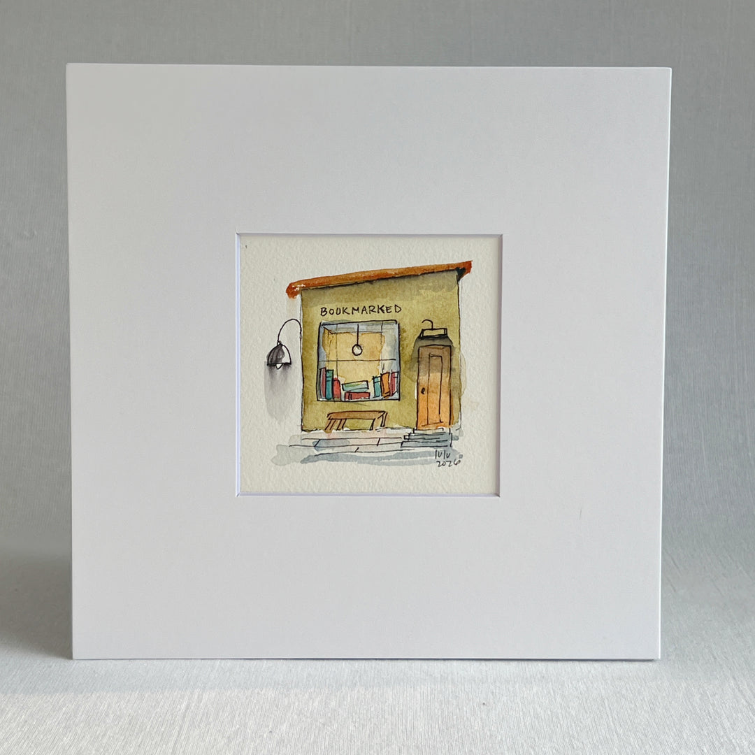 A matted watercolor painting of a cozy bookshop storefront with the word 'BOOKMARKED' above a window filled with books, a bench, a door, and a hanging lamp, shown on a white mat and white frame.