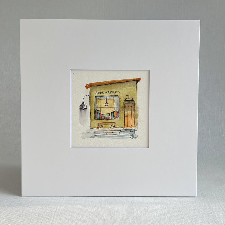 A matted watercolor painting of a cozy bookshop storefront with the word 'BOOKMARKED' above a window filled with books, a bench, a door, and a hanging lamp, shown on a white mat and white frame.