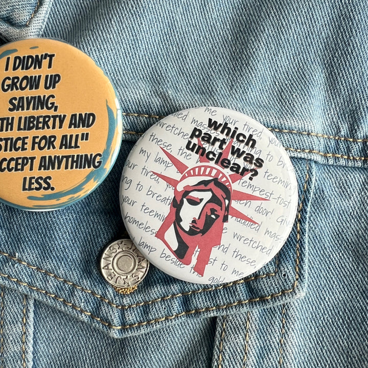 round pinback buttons pinned to a jean jacket. one with the words, 'which part was unclear?' and the face of the statue of liberty in red and blue and the other with a yellow background and the words,'I didn't grow up saying."with liberty and justice for all" to accept anything less."