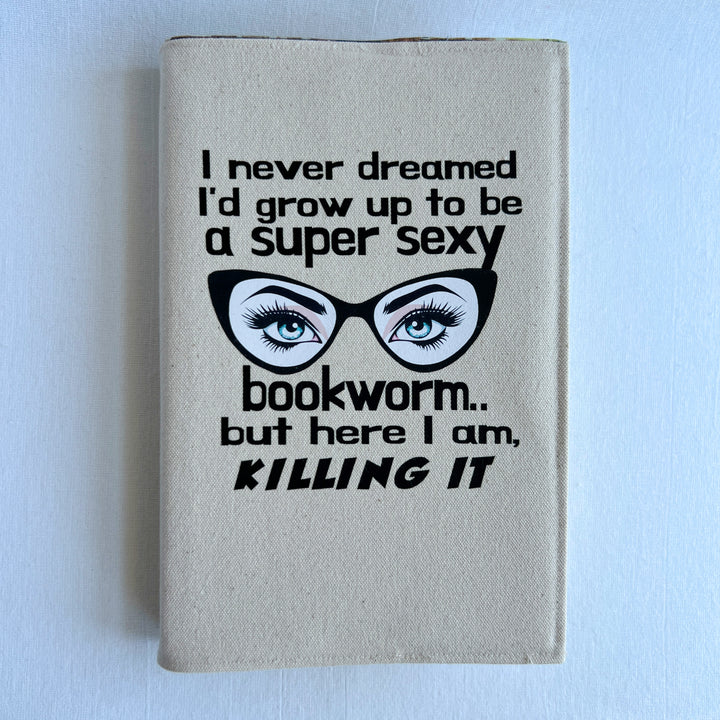 a book covered in a reusable linen-color canvas book cover, with the words, 'I never dreamed I'd grow up to be a super sexy bookworm, but here I am killing it.' A pair of eyes with long lashes behind cat eye glasses completes the design.