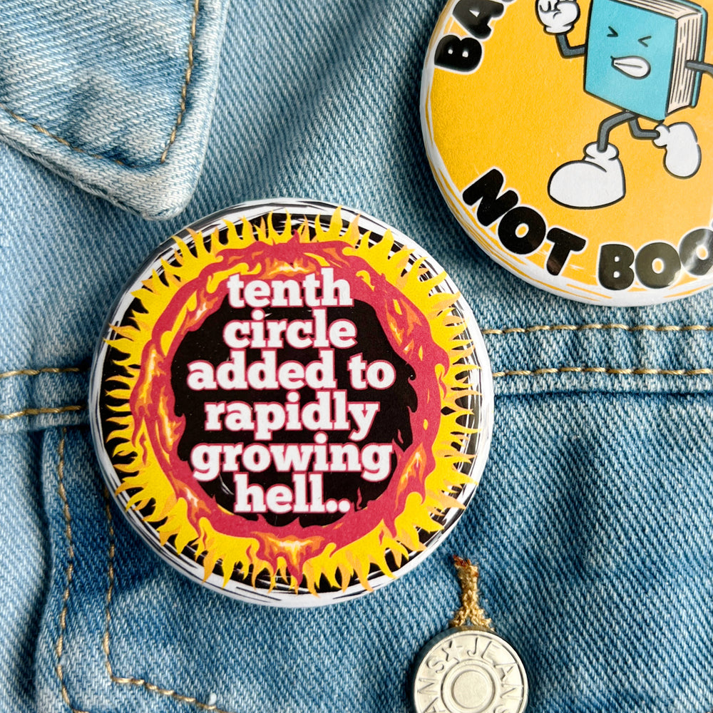 round pinback button with a black background, yellow and red flames surrounding the words, "tenth circle added to rapidly growing hell." pinned to a jean jacket for display.
