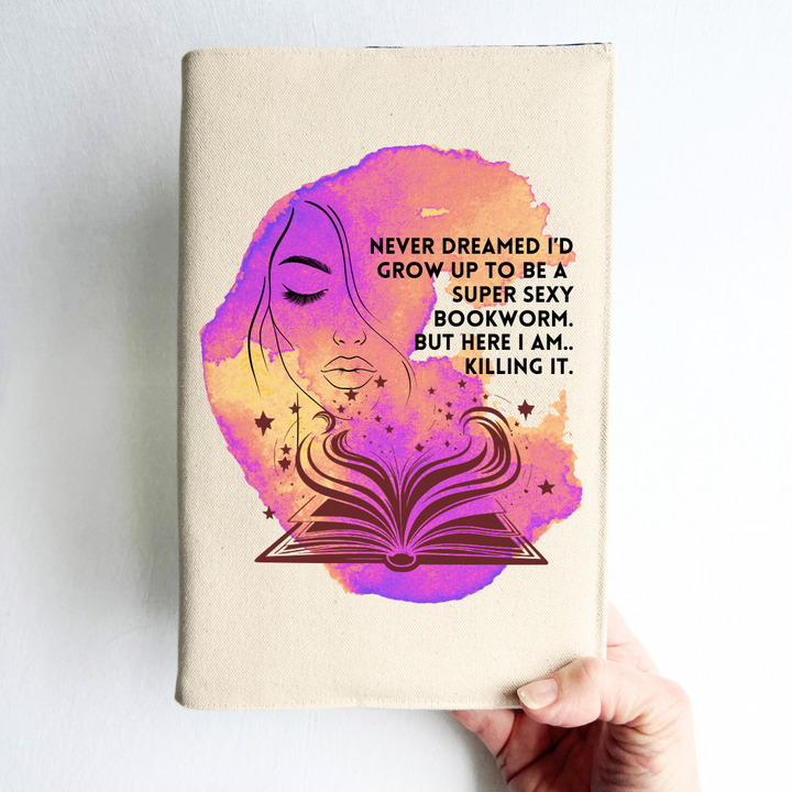 a hand holding a book covered with a linen-colored canvas with a vibrant violet golden orange watercolor swash, featuring the outline of a face with long lashes and lush lips, an open book with stars and flourishes, and the words, 'never dreamed I'd grow up to be a super sexy bookworm. but here I am, killing it.' 
