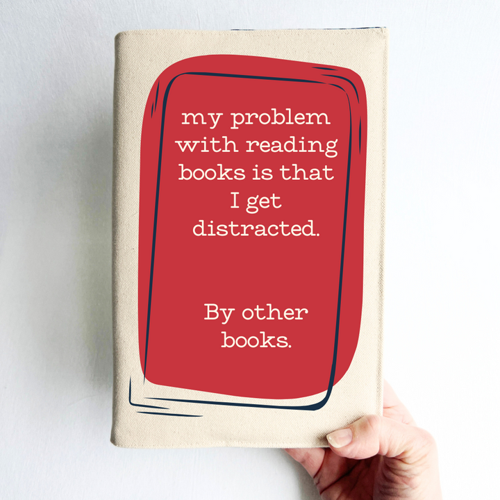 a hand holding a book covered with a linen-colored canvas reusable book cover.  A rectangular design in cherry red tops the cover, with the words, 'my problem with reading is that I get distracted.  By other books.'