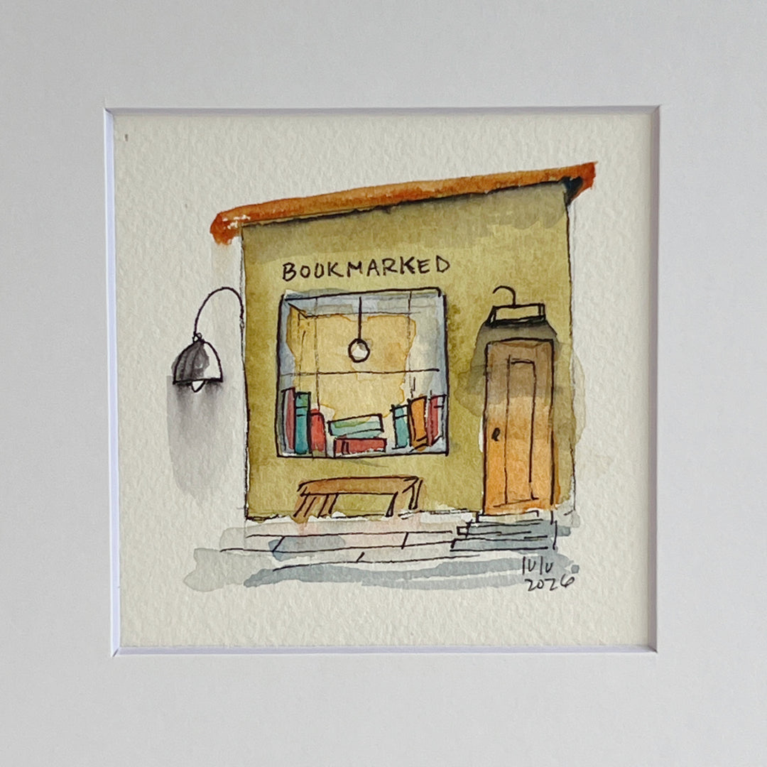 A matted watercolor painting of a cozy bookshop storefront with the word 'BOOKMARKED' above a window filled with books, a bench, a door, and a hanging lamp, shown on a white mat and white frame.