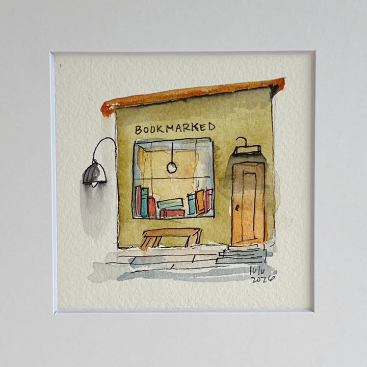 A matted watercolor painting of a cozy bookshop storefront with the word 'BOOKMARKED' above a window filled with books, a bench, a door, and a hanging lamp, shown on a white mat and white frame.
