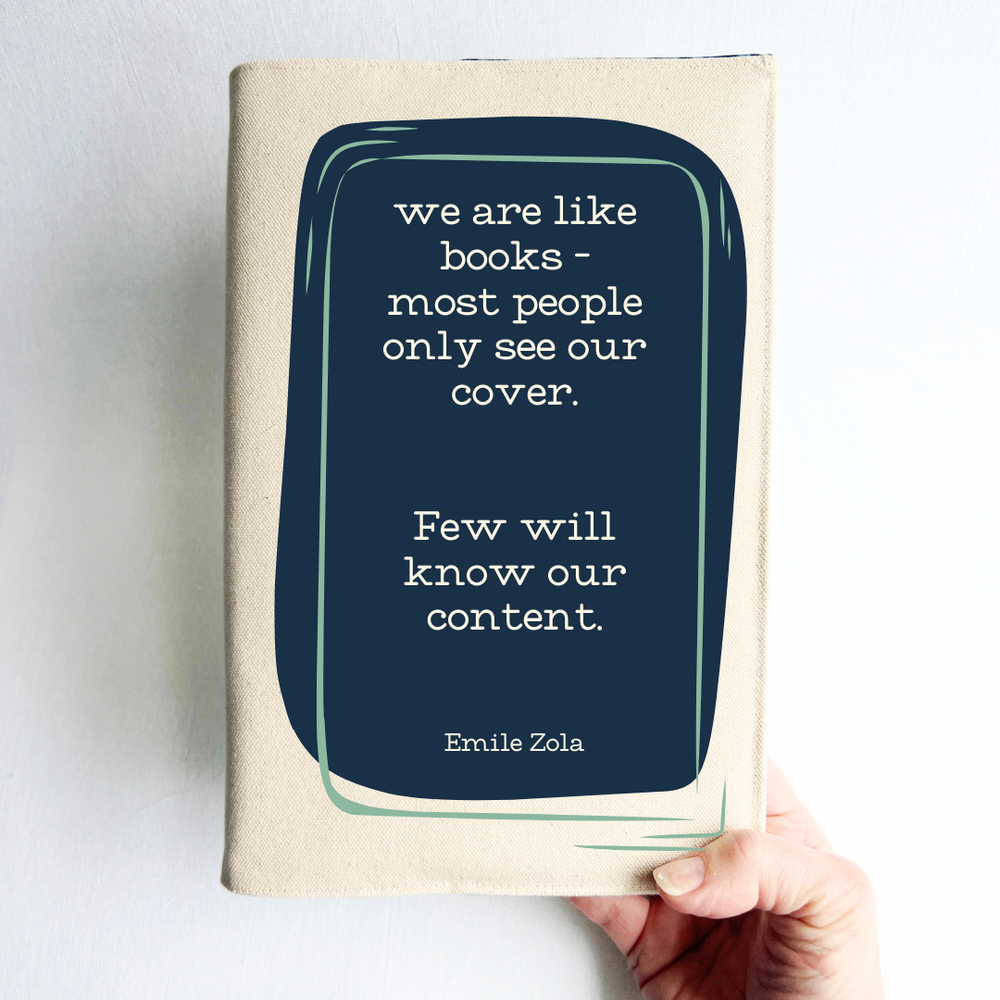 a hand holding a book covered with a linen-colored canvas reusable book cover.  A rectangular design in navy tops the cover, with the words, 'we are like books - most people only see our cover.  Few will know our content.' a quote by Emile Zola, done in white lettering.