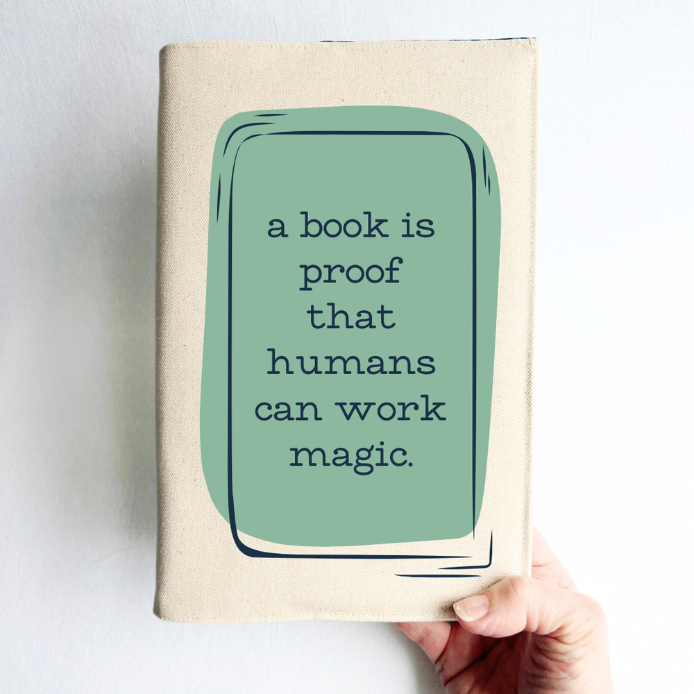 a hand holding a book covered with a linen-colored canvas reusable book cover. A rectangular design in sage green is featured in the center of the cover, with the words, 'a book is proof that humans can work magic.'