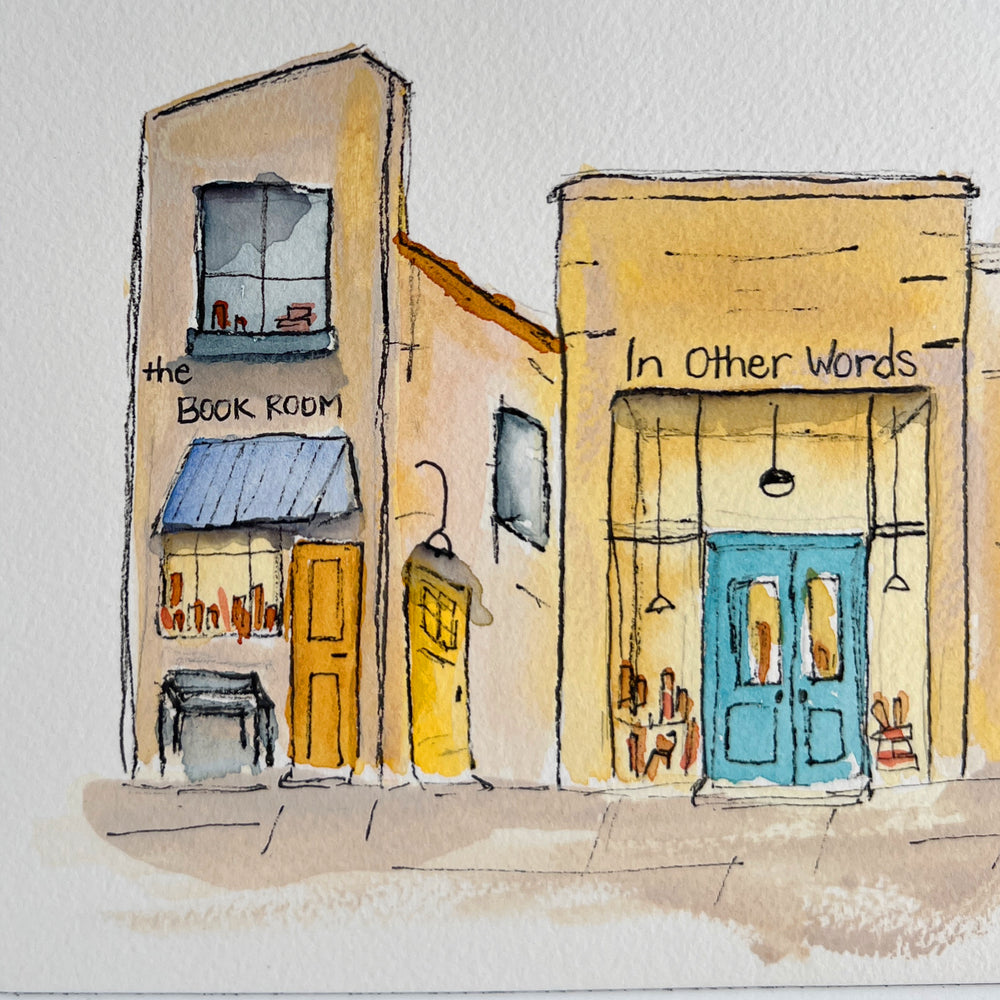 A watercolor art print depicting four stone bookstore buildings, presented with a wide white mat board against a neutral gray background.