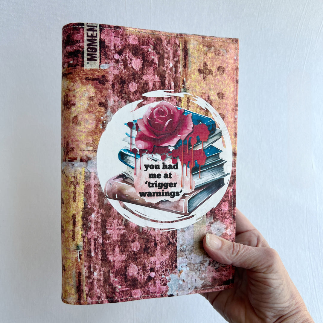 a hand holding a book covered in a colorful fabric reusable book cover in colors of pinks and orange. A white circle on the front contains an original design with a stack of books, a rose, blood drips and the words, 'you had me at trigger warnings' finishes the look.  Meant for readers of dark romance.