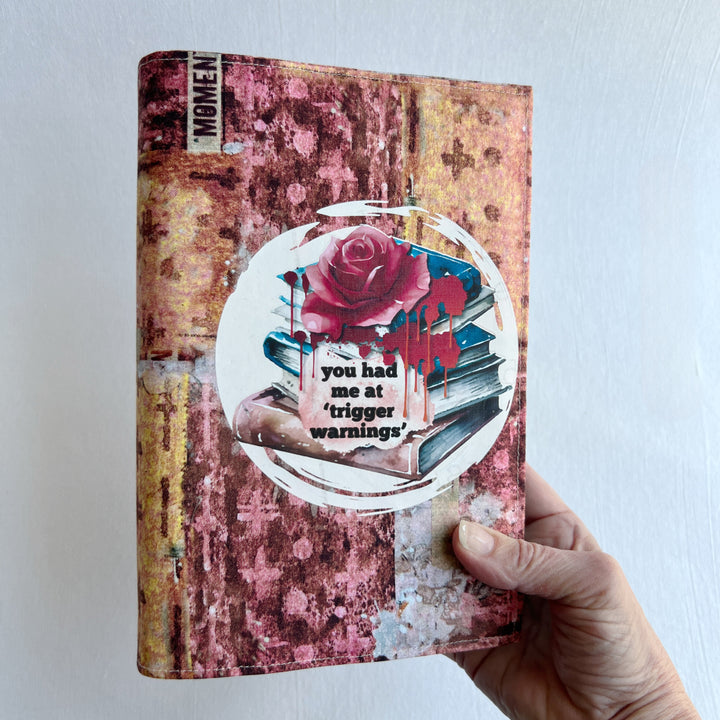 a hand holding a book covered in a colorful fabric reusable book cover in colors of pinks and orange. A white circle on the front contains an original design with a stack of books, a rose, blood drips and the words, 'you had me at trigger warnings' finishes the look.  Meant for readers of dark romance.