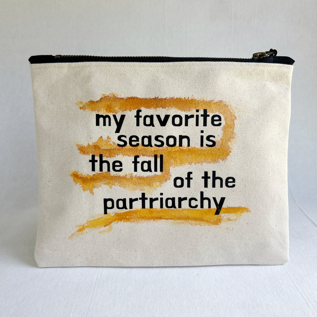 An off-white canvas zipper pouch with a black zipper and a wooden tassel. The front features a black printed text reading 'my favorite season is the fall of the partriarchy' overlaid on top of three horizontal orange-brown watercolor brushstrokes.
