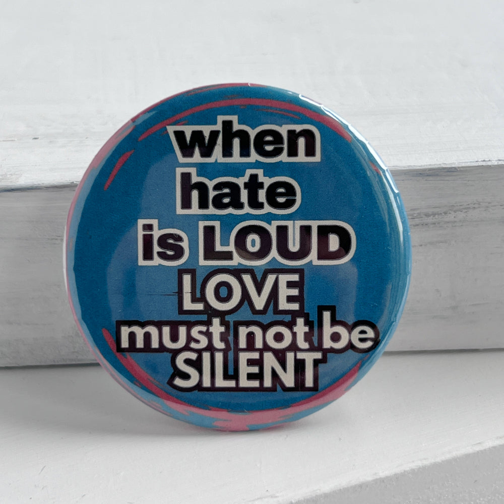 a round pinback button with a blue background and streaks of read, with the words, "when hate is LOUD, LOVE must not be SILENT."