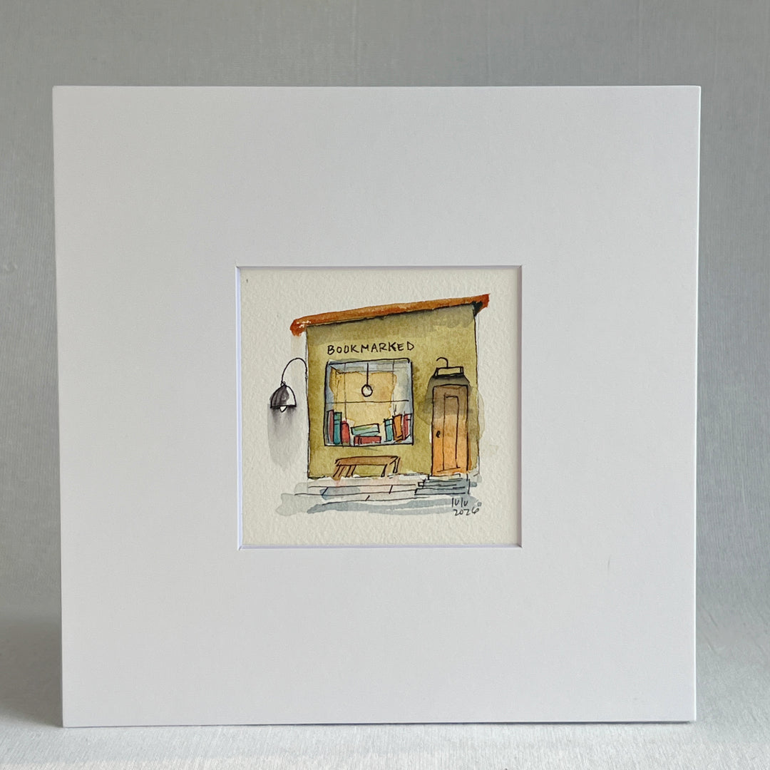 A matted watercolor painting of a cozy bookshop storefront with the word 'BOOKMARKED' above a window filled with books, a bench, a door, and a hanging lamp, shown on a white mat and white frame.