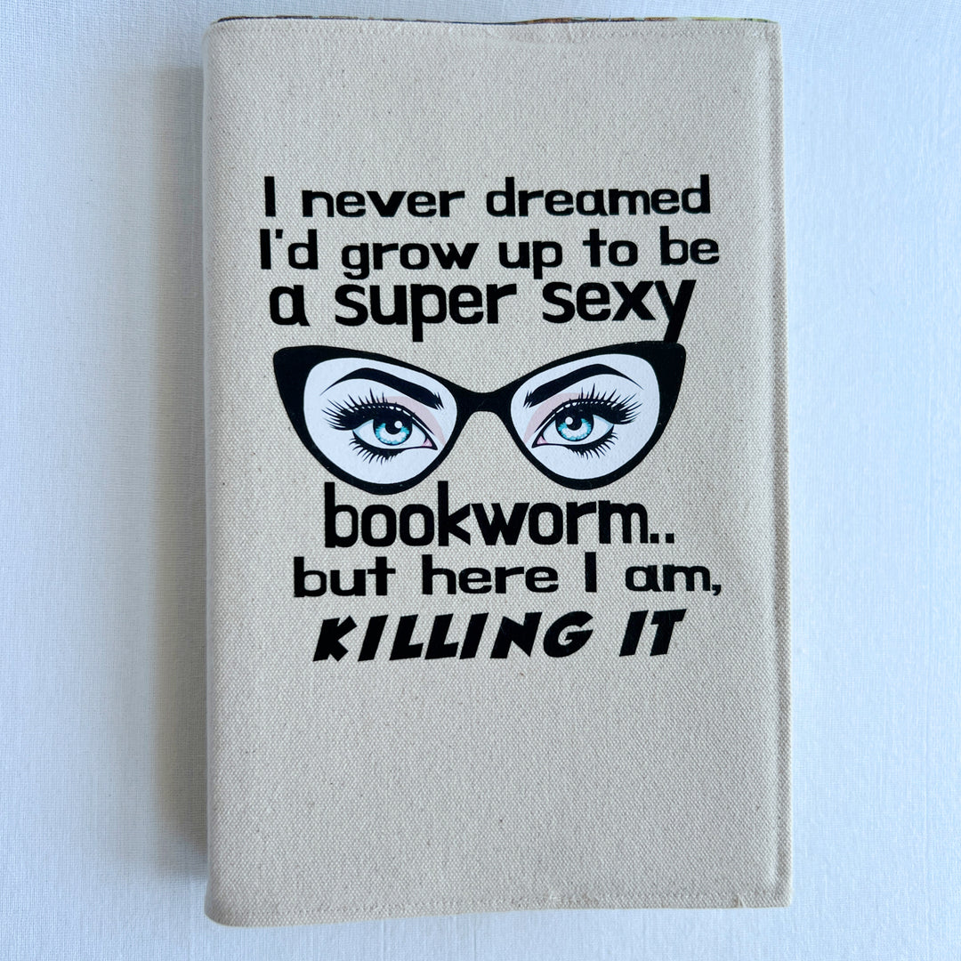 a book covered in a reusable linen-color canvas book cover, with the words, 'I never dreamed I'd grow up to be a super sexy bookworm, but here I am killing it.' A pair of eyes with long lashes behind cat eye glasses completes the design.