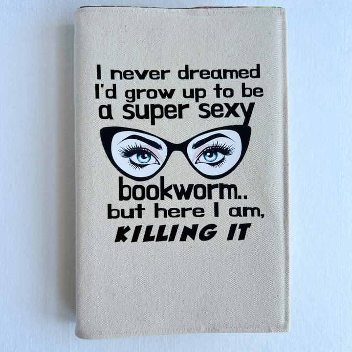 a book covered in a reusable linen-color canvas book cover, with the words, 'I never dreamed I'd grow up to be a super sexy bookworm, but here I am killing it.' A pair of eyes with long lashes behind cat eye glasses completes the design.