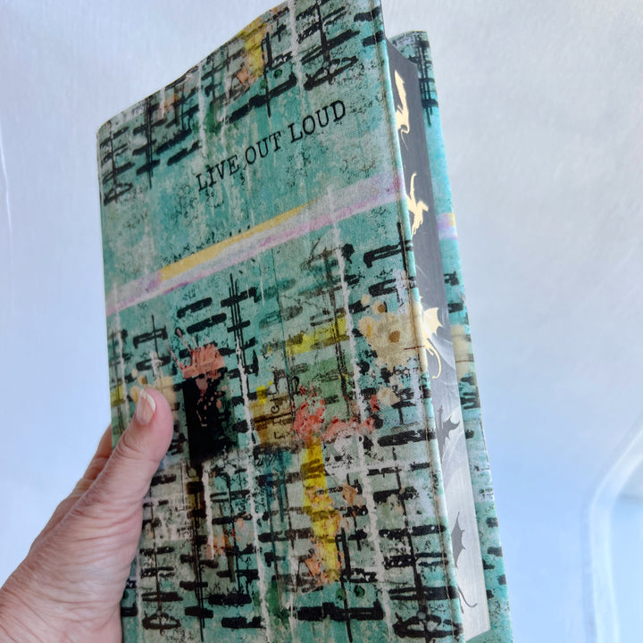 a hand holding a book covered in a vibrant blue green fabric reusable book cover with the words, 'live out loud' printed on the top.